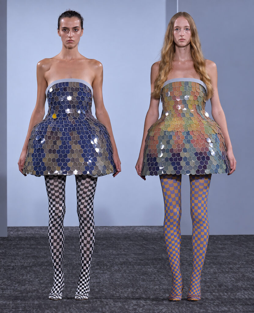 Two models wear strapless scale-patterned bell-shaped dresses with checkered patterned tights on a runway.