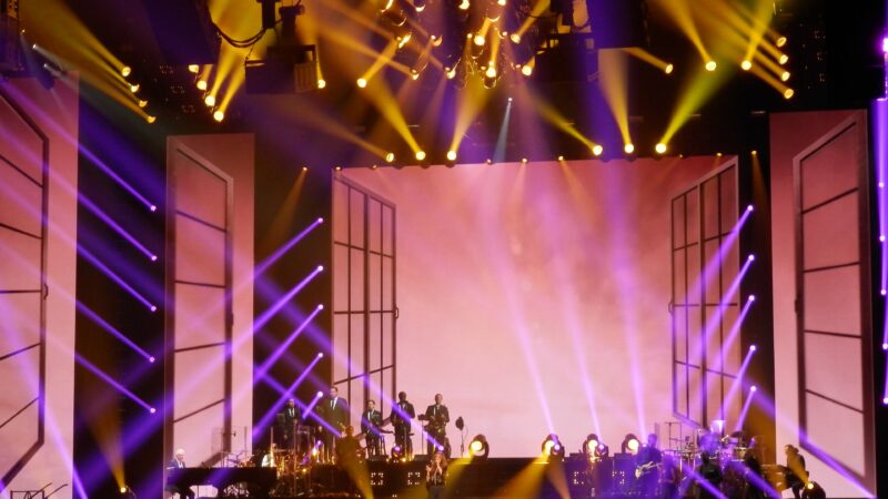 A concert stage with purple and gold lighting, a band, and a singer performing in front of a large screen and crowd.