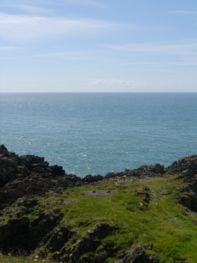 A grassy rocky headland overlooking the blue sea under a partly cloudy sky.