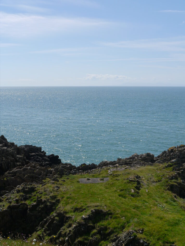 A grassy rocky headland overlooking the blue sea under a partly cloudy sky.
