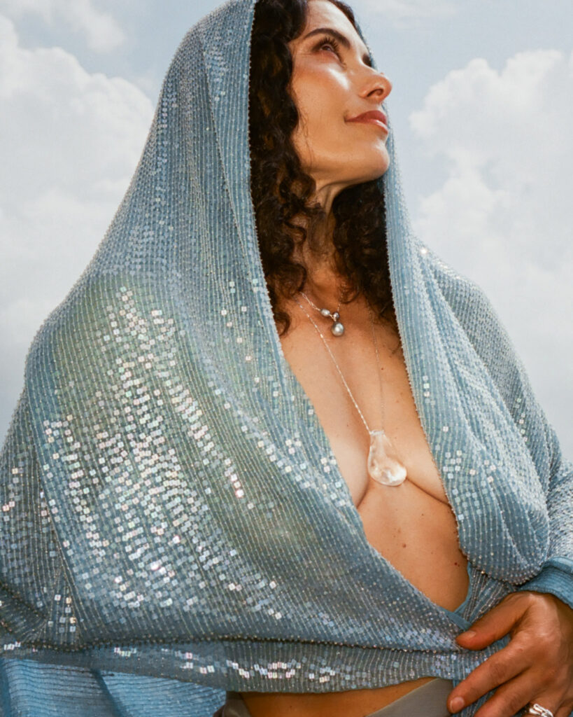 A woman in a sparkling blue sequined hooded wrap wears a layered necklace with a pearl and a teardrop crystal pendant.