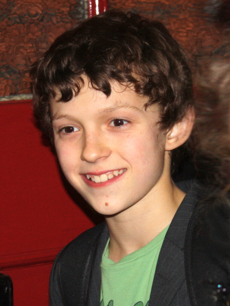 A young boy with curly brown hair smiles, wearing a green shirt and dark blazer.