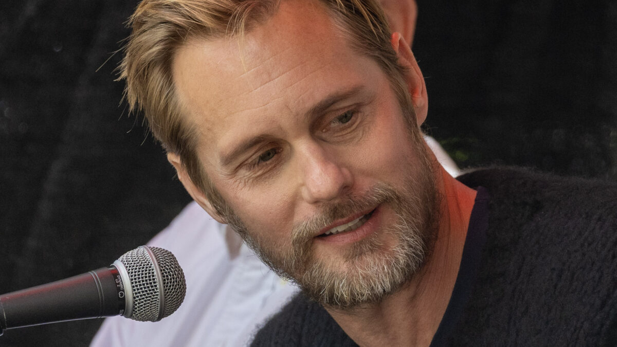 A bearded man speaking into a microphone outdoors at a public event.