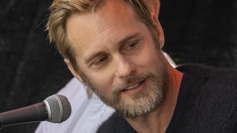 A bearded man speaking into a microphone outdoors at a public event.