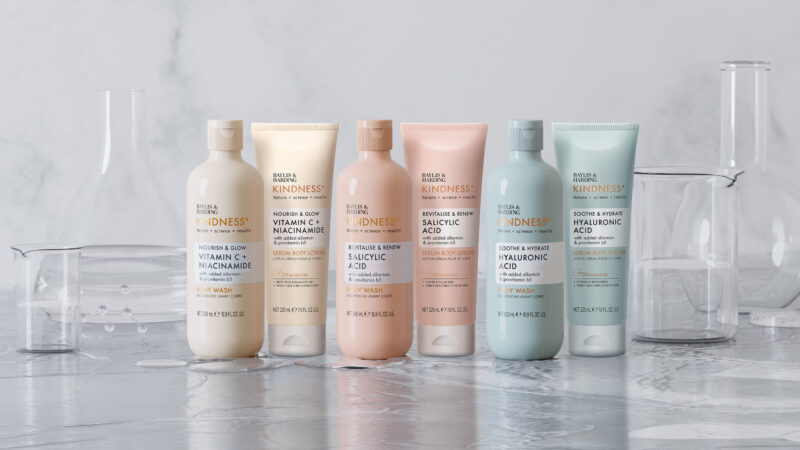 Six Baylis & Harding Kindness+ body wash and serum body lotion bottles arranged among laboratory glassware.