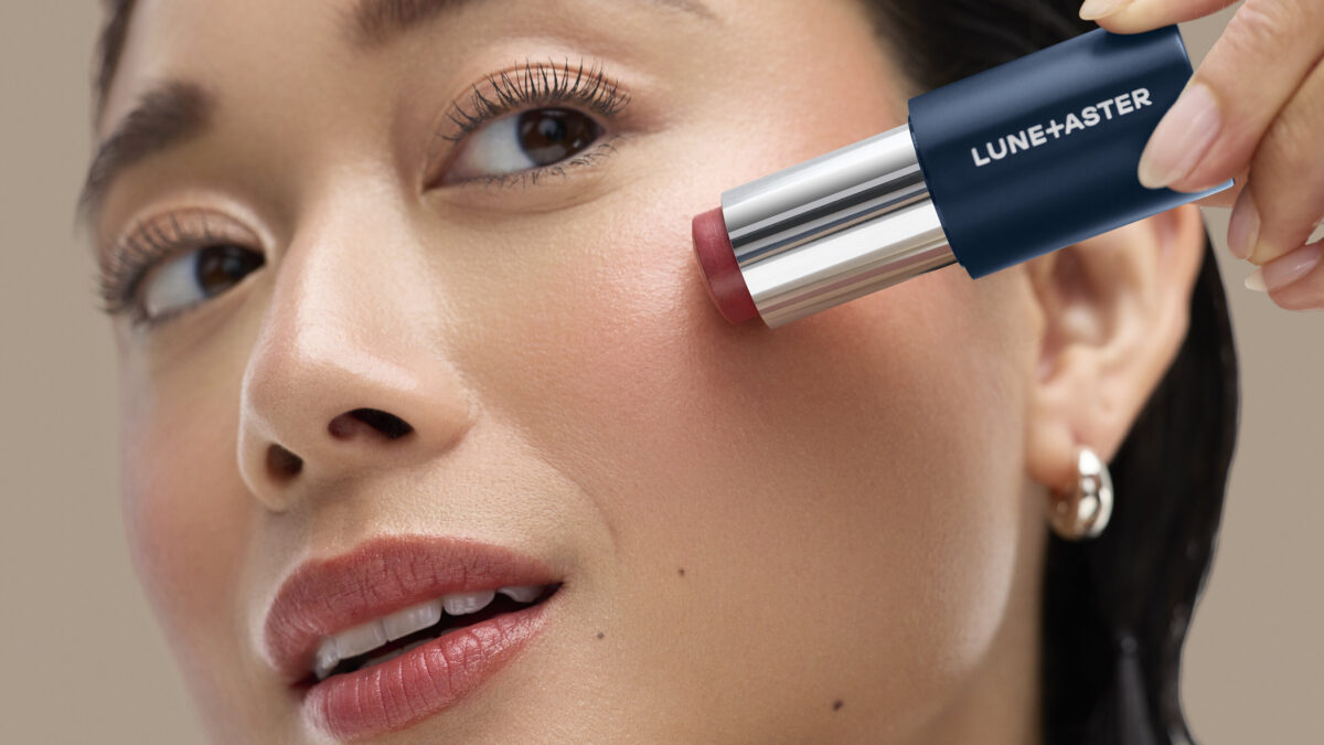Close-up of a woman applying a cream blush stick labeled Lune+Aster to her cheek.