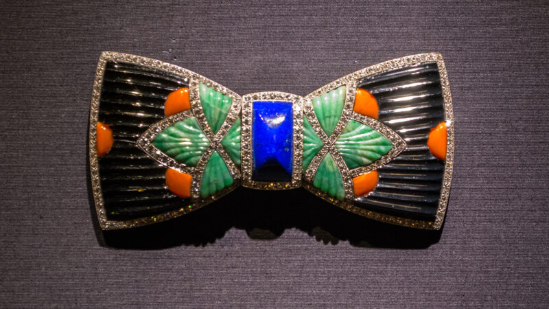 A bow-shaped Boucheron brooch with black, green, orange, and blue gemstones and diamond accents displayed on gray fabric.