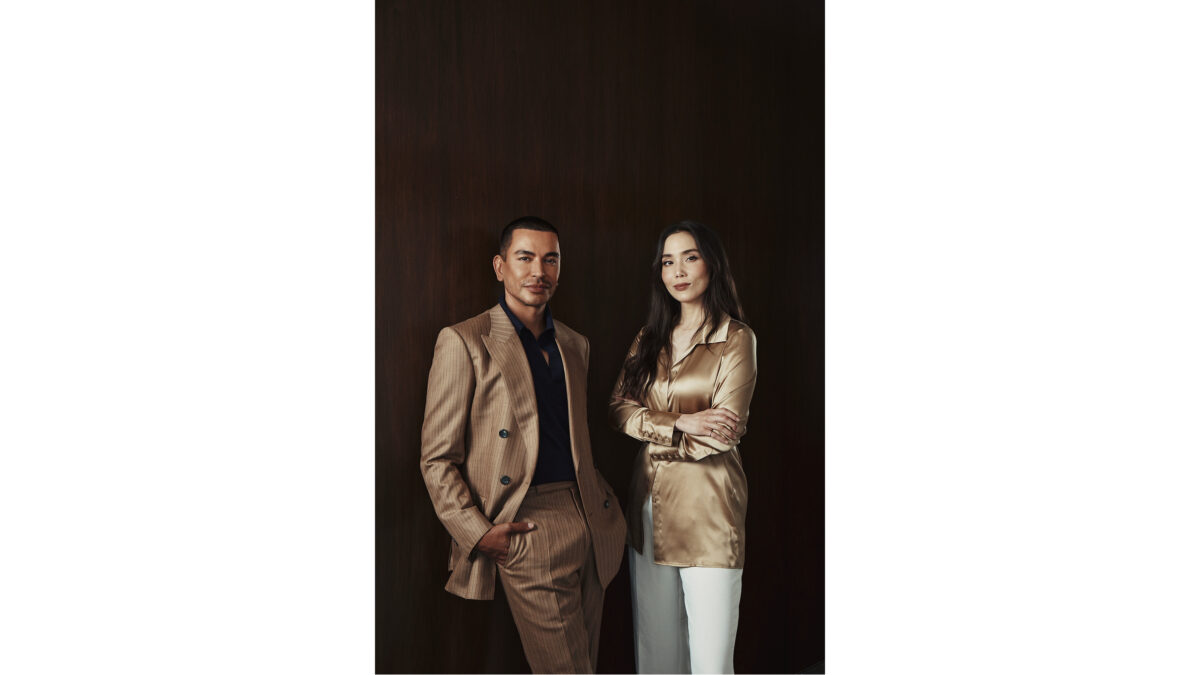 A man in a tan suit and a woman in a gold satin blouse stand together against a dark wood paneled wall.
