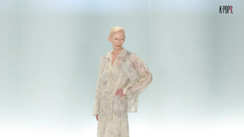 A woman with short blonde hair wears a feathery, textured Chanel outfit on a runway.