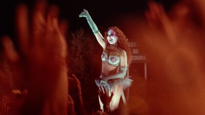 A performer in a sparkly costume and theatrical makeup raises a gloved arm on stage above an audience with raised hands.