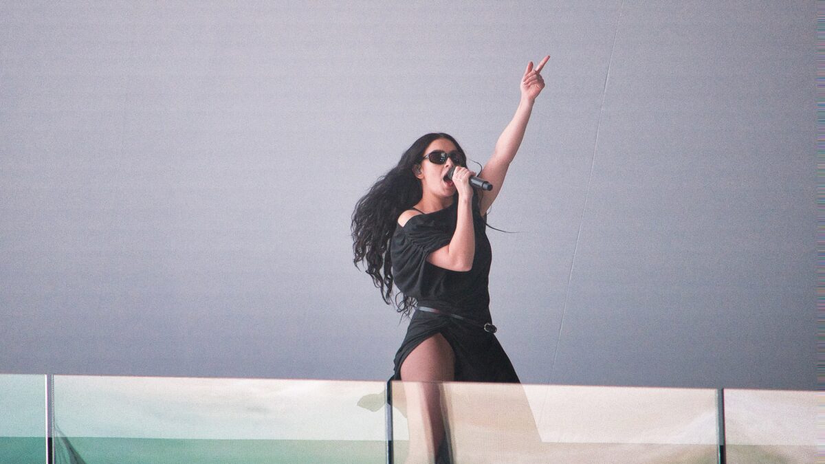 A performer in a black dress sings into a microphone while pointing upward on stage.