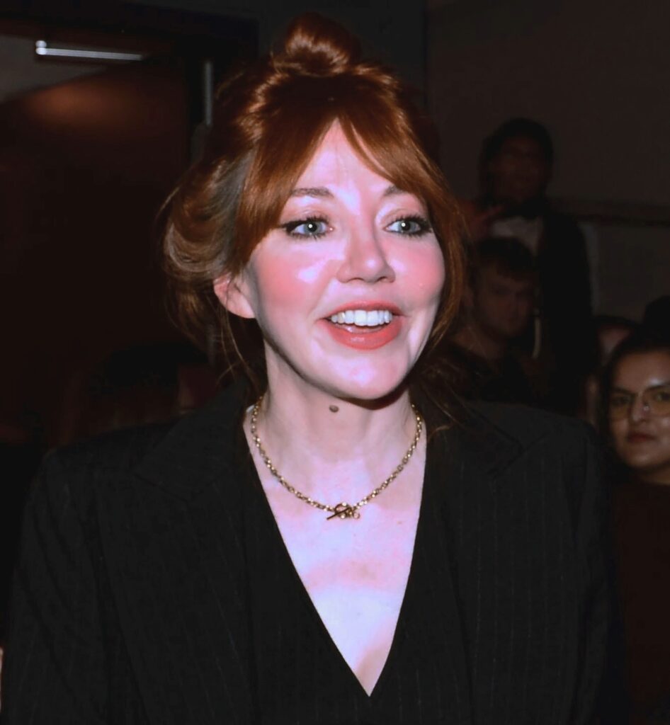 A woman with auburn hair in an updo smiles while wearing a black blazer and gold necklace at an indoor event.