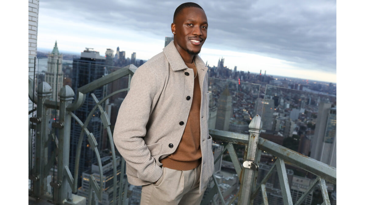 A man in a houndstooth jacket smiles on a rooftop terrace with a city skyline behind him.