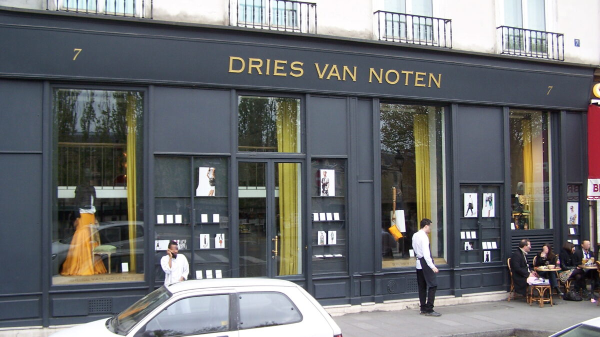 Storefront of the Dries Van Noten boutique with gold lettering, street scene with parked cars and pedestrians.