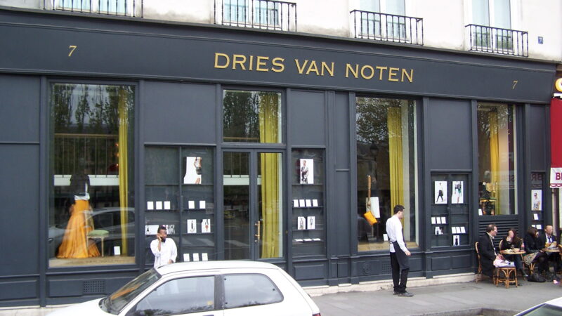 Storefront of the Dries Van Noten boutique with gold lettering, street scene with parked cars and pedestrians.