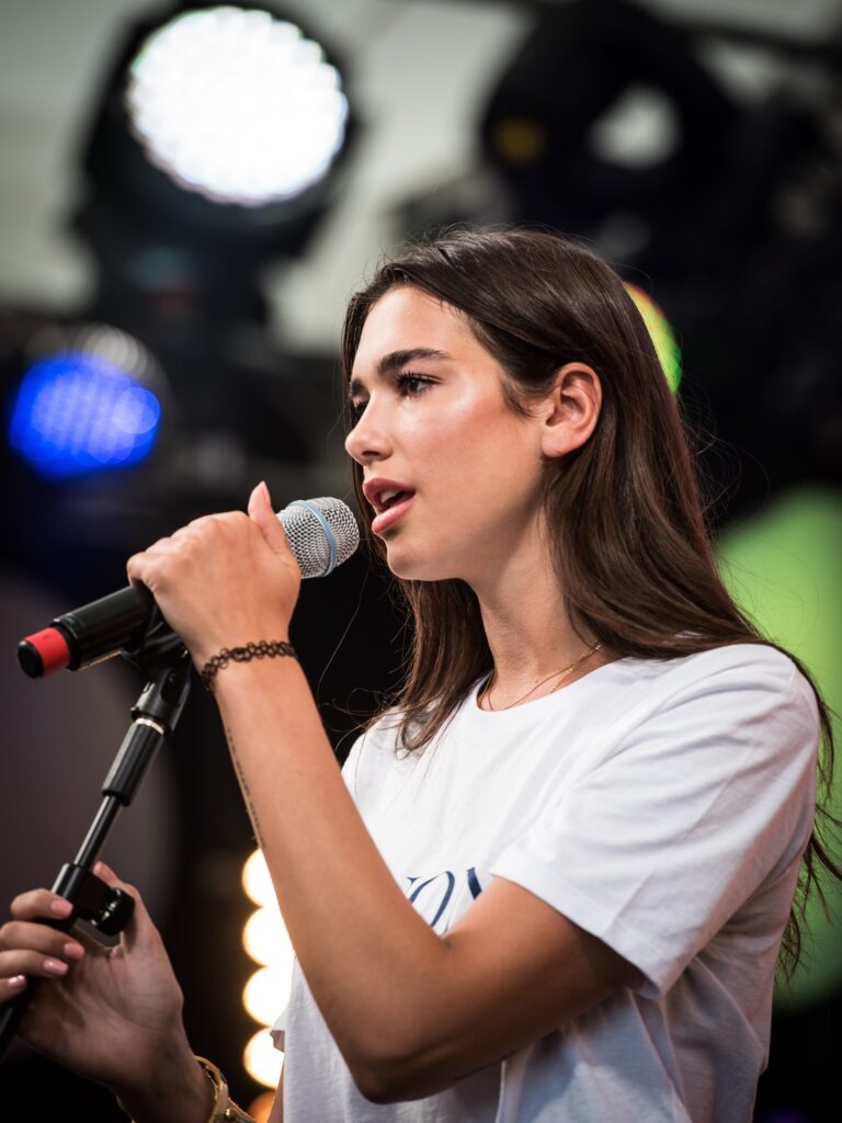A woman with long dark hair sings into a microphone on a stage with stage lights in the background.