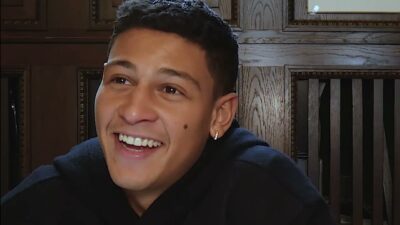 A young man with short dark hair smiles broadly while being interviewed in a wood-paneled room.
