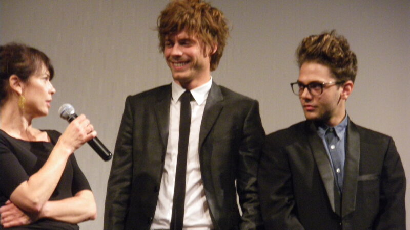 Three people on stage, a woman holding a microphone and two men in dark suits, at a film festival event.