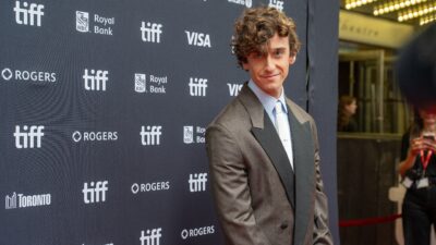 A curly-haired man in a suit smiles at the camera in front of a TIFF sponsor backdrop.
