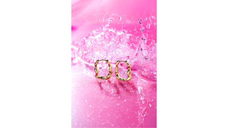 A pair of textured gold hoop earrings sit on a wet pink surface with a water splash behind them.