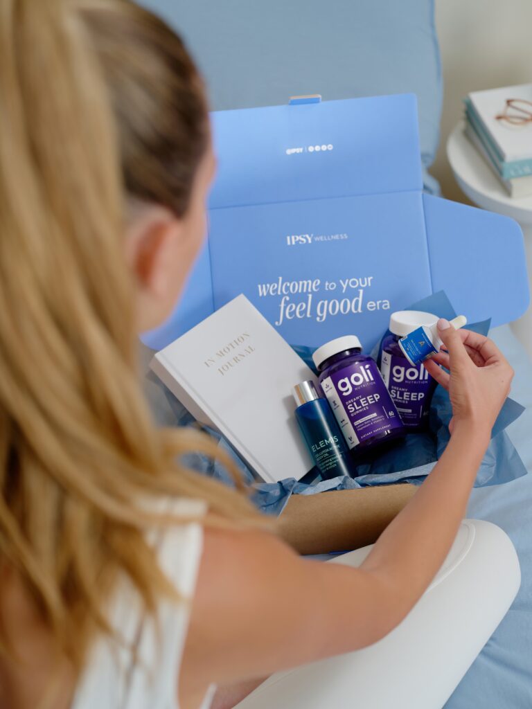 A woman unboxes an IPSY Wellness box containing a journal, body oil, and Goli sleep gummies.