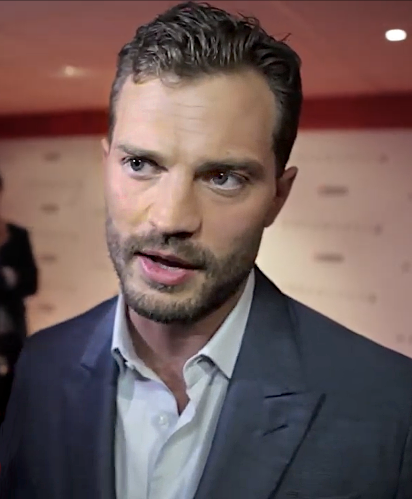 Jamie Dornan speaking in a suit at a media event in 2016.