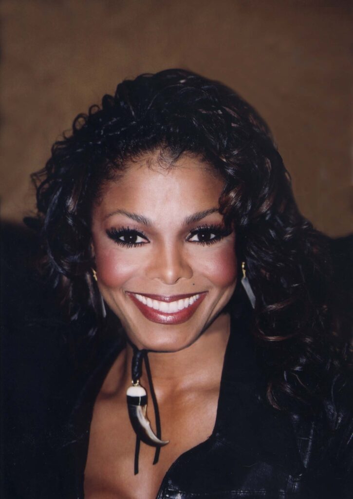 Close-up photo of a smiling woman with curly dark hair wearing a black outfit and a tooth-shaped pendant necklace.
