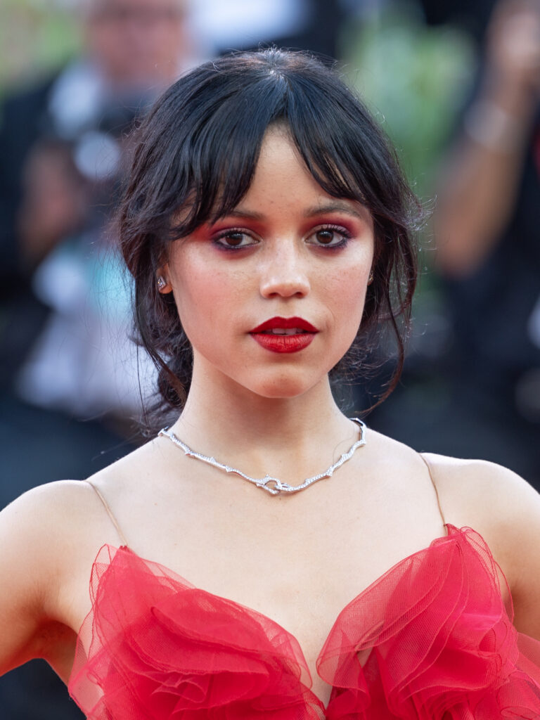 A woman with dark hair, red eyeshadow, and red lipstick wears a red ruffled dress and diamond necklace on a red carpet.