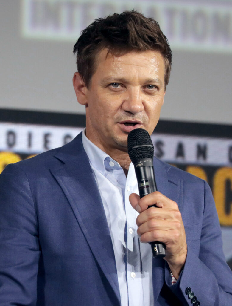 A man in a blue blazer speaks into a microphone in front of a convention backdrop.