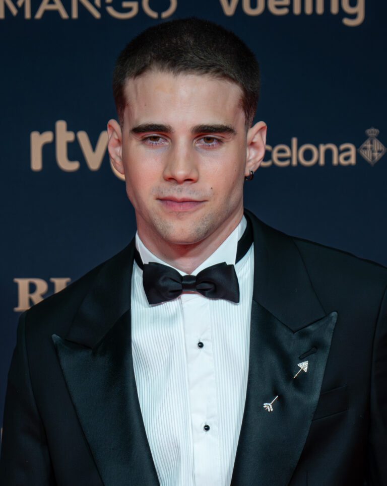 A young man in a black tuxedo with a bow tie poses at a red carpet event with sponsor logos behind him.