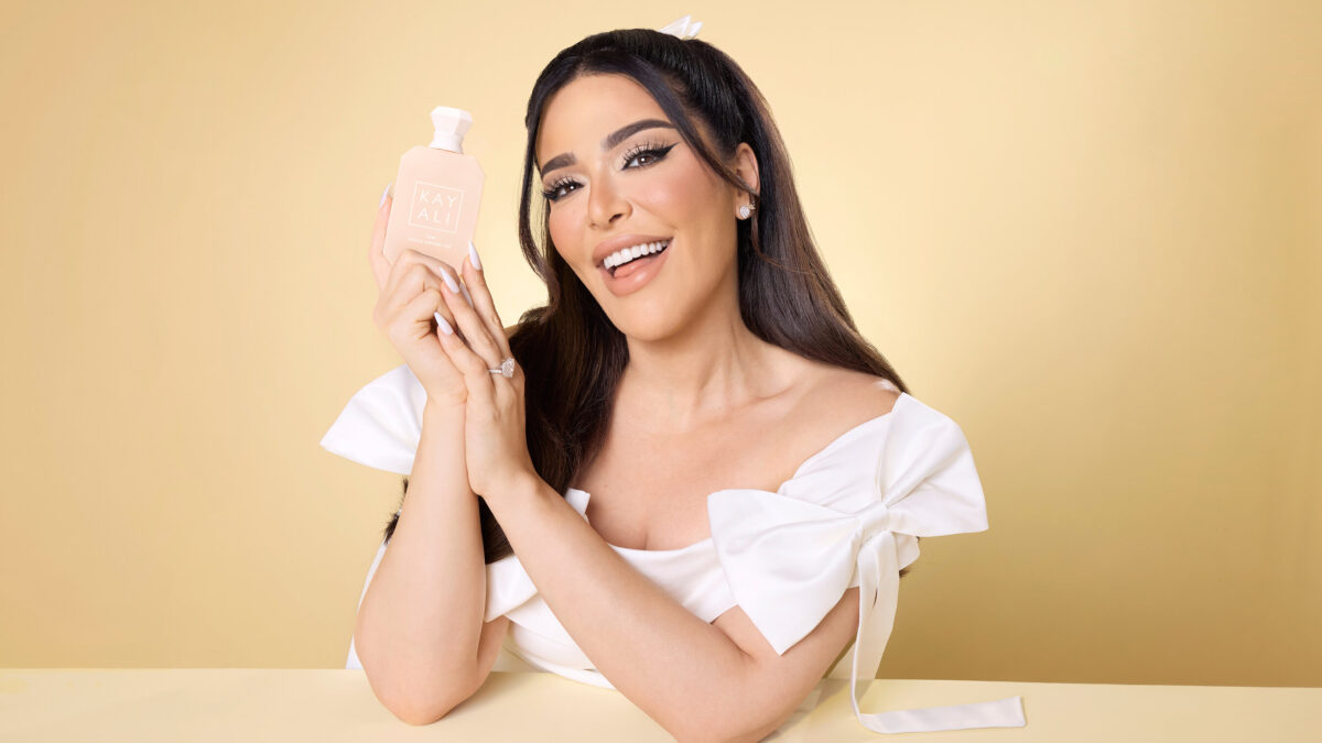 A smiling woman in a white top holds a pink Kayali perfume bottle against a tan background.