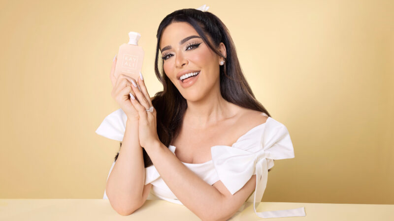 A smiling woman in a white top holds a pink Kayali perfume bottle against a tan background.