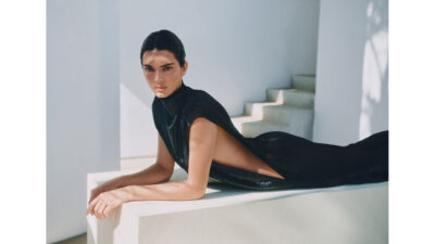 A woman with dark hair wears a black gown while reclining on a white ledge near a staircase.
