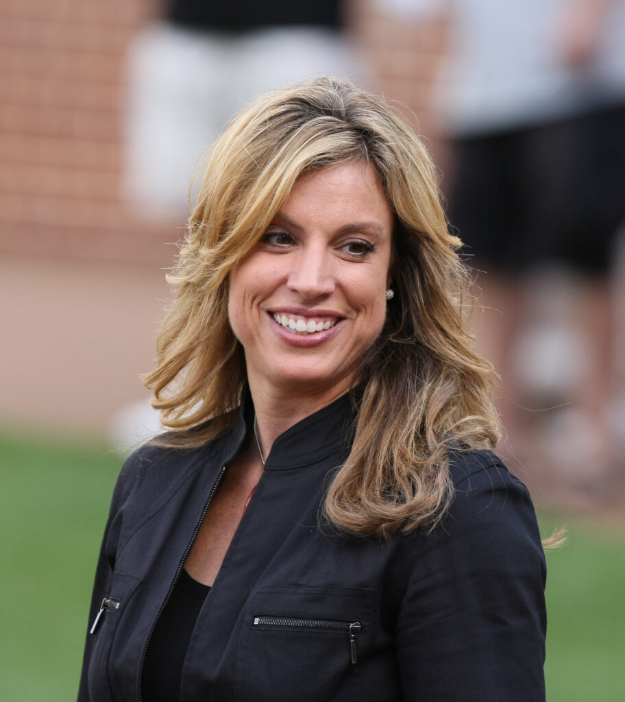 A smiling woman with blonde hair wearing a black jacket stands outdoors near a brick building.