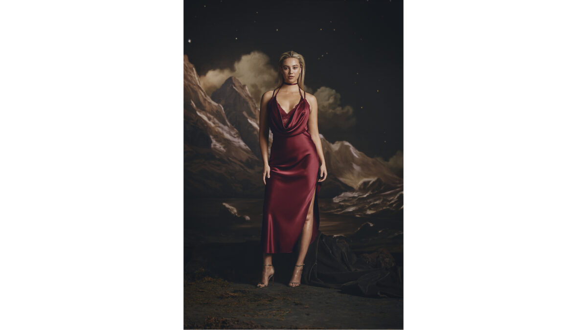 A woman in a dark red satin slip dress stands before a painted backdrop of mountains and a starry sky.