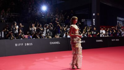 A woman in a sheer floral gown walks a red carpet in front of photographers at a BIFF 2025 event.