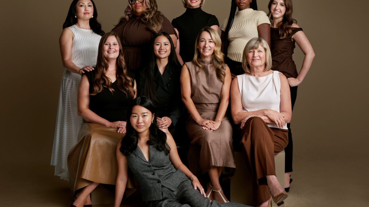 Group studio portrait of ten women of varying ages and ethnicities posing together in neutral-toned outfits.