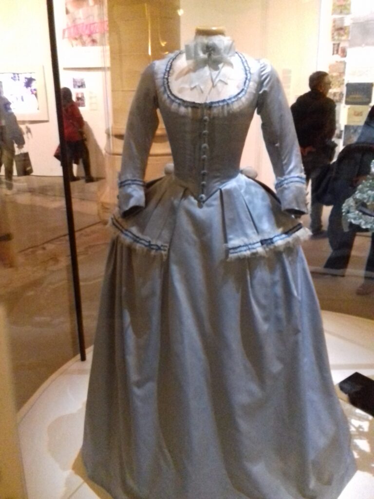 A pale blue silk 18th-century style travel costume displayed on a mannequin in a museum case.