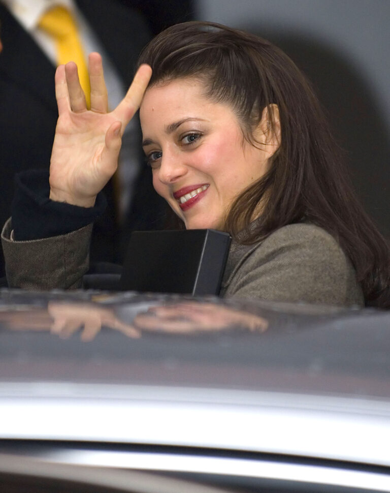 A woman with dark hair waves while smiling, seen through the open roof of a car.