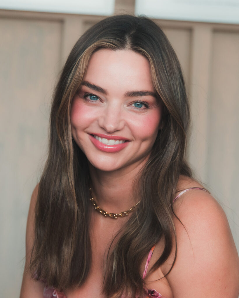 A smiling woman with long brown hair and a gold necklace poses for a close-up photo outdoors.