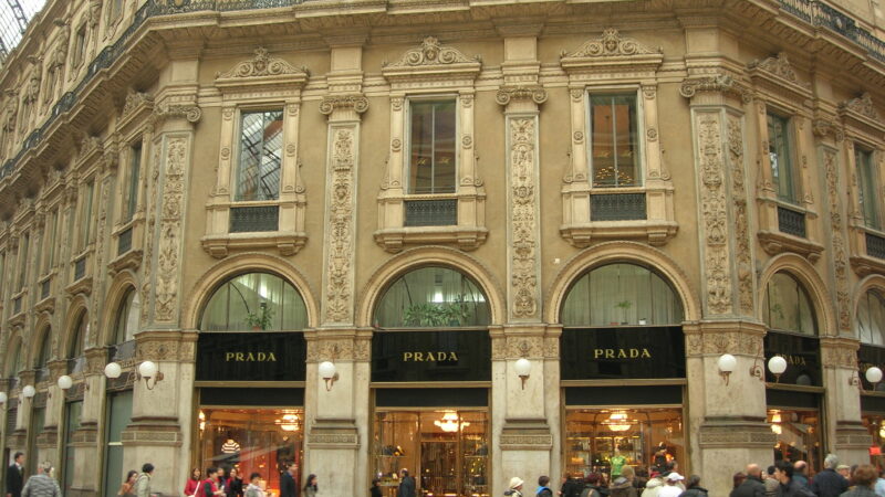 Prada store signage on an ornate historic building facade in Milan with pedestrians walking by.