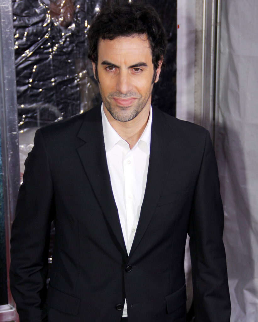 A man with curly dark hair and stubble wears a black suit and white shirt at an event.