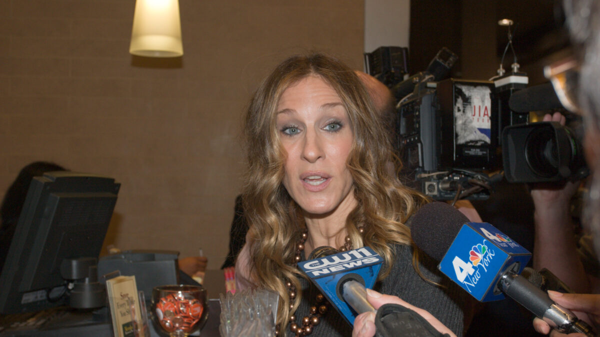 A woman with long wavy hair is interviewed by reporters holding CW11 and NBC New York microphones.