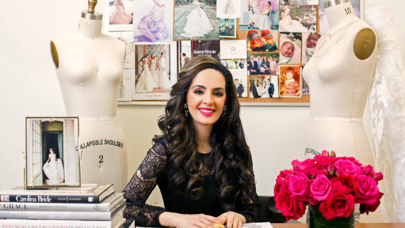 Sareh Nouri sits at a desk sketching, surrounded by dress forms, a mood board, and pink roses.