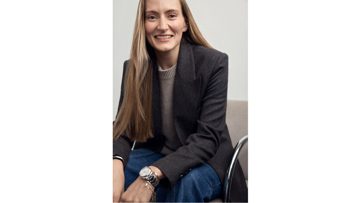 A smiling woman with long hair wearing a dark blazer sits on a chair for a portrait.