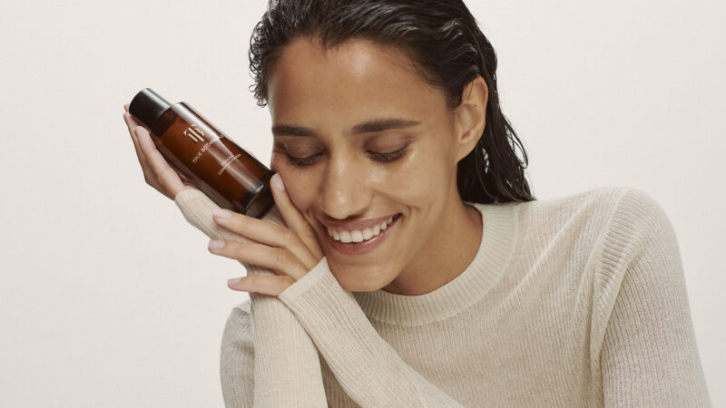 A smiling woman in a cream sweater holds a brown True Botanicals bottle against her cheek.