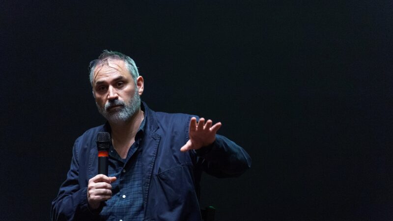 A bearded man in a dark jacket holds a microphone and gestures while speaking on a dimly lit stage.